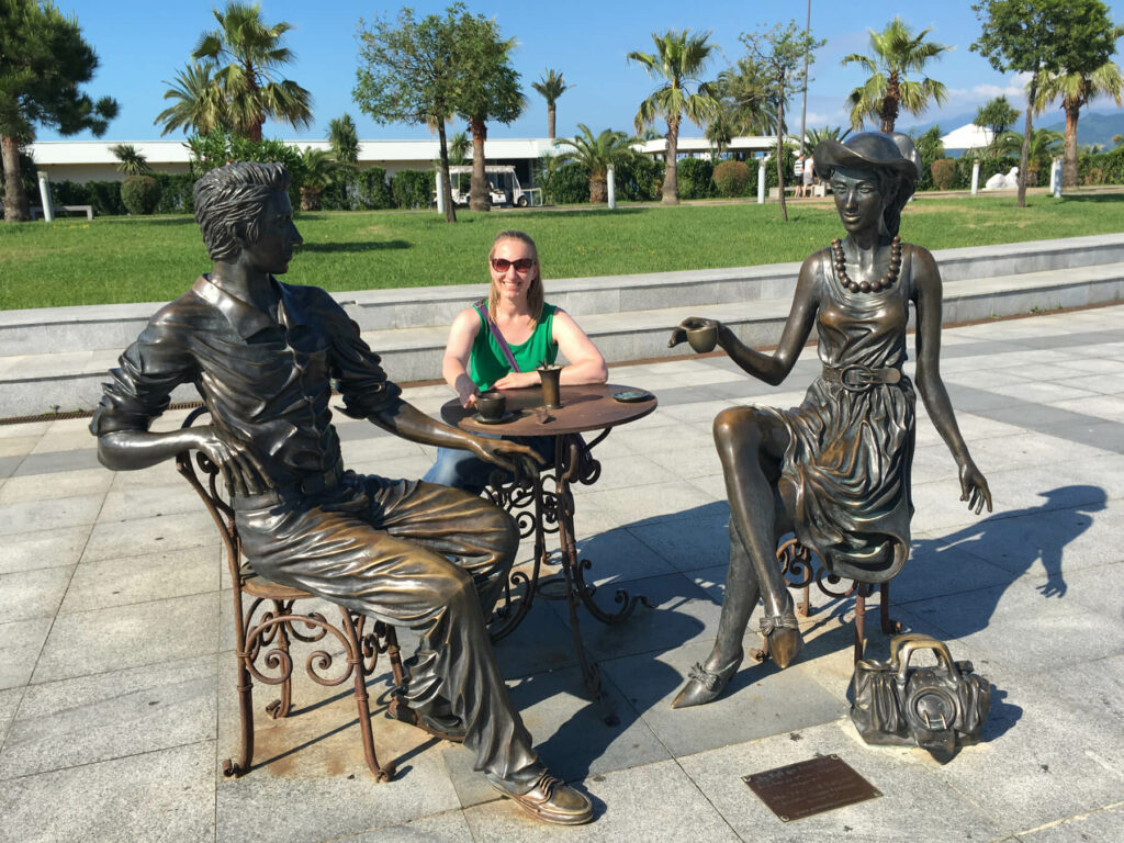 One female figure at a table with two statues as if in conversation with palm trees in the background.